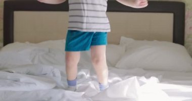 Kıvırcık saçlı, tişörtlü ve şortlu bir çocuk. Sabahları yatakta zıplıyor..