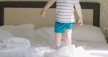 Kıvırcık saçlı, tişörtlü ve şortlu bir çocuk. Sabahları yatakta zıplıyor..