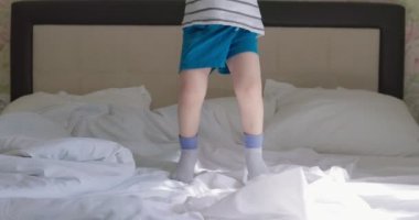 Kıvırcık saçlı, tişörtlü ve şortlu bir çocuk. Sabahları yatakta zıplıyor..