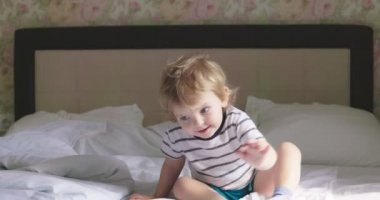 Kıvırcık saçlı, tişörtlü ve şortlu bir çocuk. Sabahları yatakta zıplıyor..