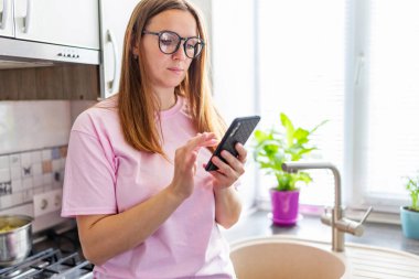 Beautiful young woman in eyeglasses using mobile phone at home kitchen. Brunette girl reading or typing a text message.