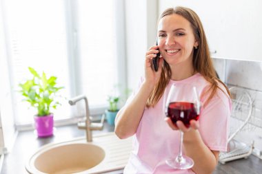 Mutfakta elinde bir bardak kırmızı şarapla duran ve akıllı telefonunu kullanan mutlu ve rahat genç bir kadın..