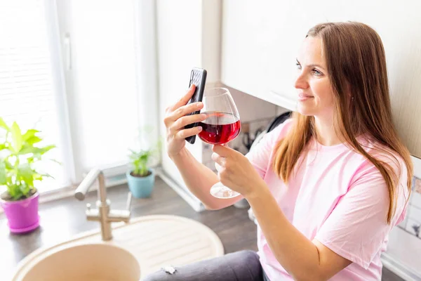 Happy relaxed young woman standing in kitchen with glass of red wine and using her smartphone.