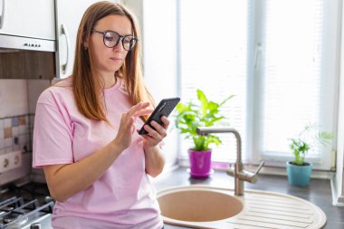Beautiful young woman in eyeglasses using mobile phone at home kitchen. Brunette girl reading or typing a text message.