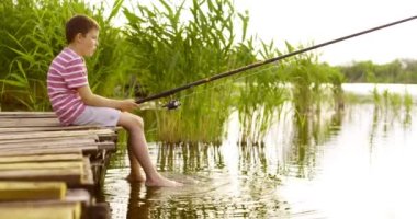 Little boy sitting on the edge of a wooden dock and fishing in lake at sunset. Summer outdoor leisure activity