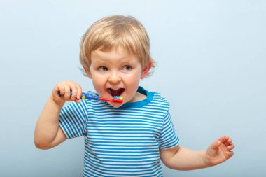 Little blonde boy brushing teeth with toothbrush on blue background. Dental hygiene.