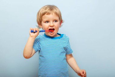 Little blonde boy brushing teeth with toothbrush on blue background. Dental hygiene.