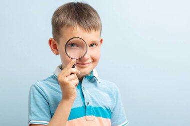 Curious schoolboy in casual clothes looking through the magnifying glass on blue background.