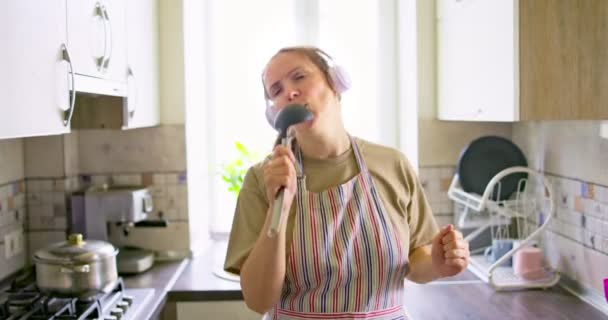 在厨房做饭的时候 耳机里的漂亮积极的女人一边听着音乐 一边像话筒一样对着小勺唱歌 在厨房放轻松 — 图库视频影像