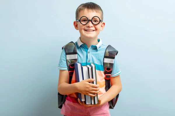 Happy little boy in glasses with backpack hold books on blue background. Childhood lifestyle concept. Education in school.