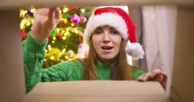 Beautiful young woman in a Christmas hat opening Christmas gift box with excited surprised face feeling happy on New Year's Eve sitting at home near glowing christmas tree. Magic moments