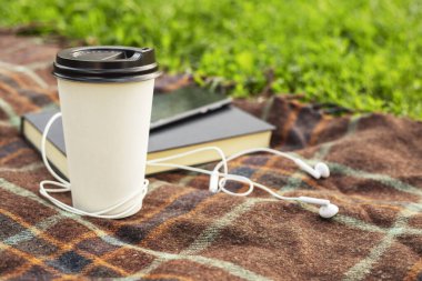 Hobby, relaxation and recreation concept. White cup of hot coffee with headphones, smartphone and book in garden with copy space.