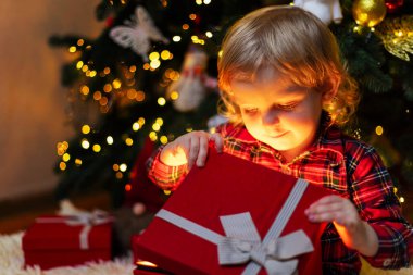 Cute little boy opening magic Christmas gift by a Christmas tree in cozy living room in winter. Merry Christmas!