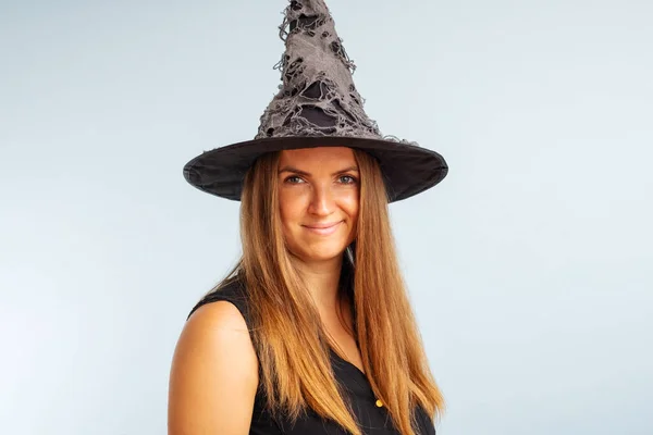 Happy Halloween! Happy brunette woman in halloween witch costume with black hat on a light background.