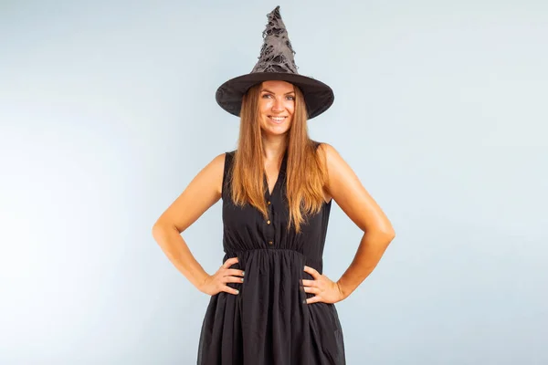 Happy Halloween! Happy brunette woman in halloween witch costume with black hat on a light background.