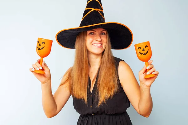 Happy Halloween! Happy brunette woman in halloween witch costume with black hat holding glasses of cocktails on a light background. Halloween party.