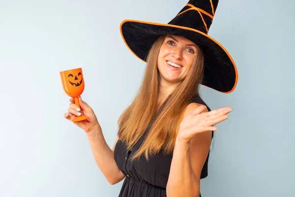 Happy Halloween! Happy brunette woman in halloween witch costume with black hat holding glass of cocktail on a light background. Halloween party.