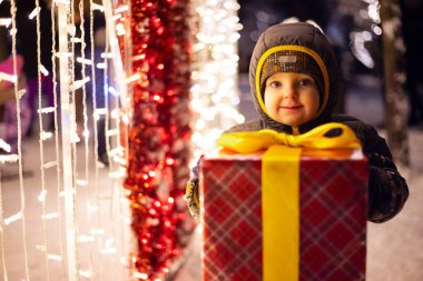 Noel Baba şapkalı mutlu çocuk kış akşamları parlayan ampullerin yanında Noel hediyesini paketliyor. Noel ve Yeni Yıl kavramı.