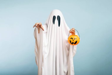 Cadılar Bayramı Konsepti. Siyah gözlü beyaz bir hayalet, mavi arka planda şeker için balkabağı sepetinden yapılmış bir yatak örtüsü..