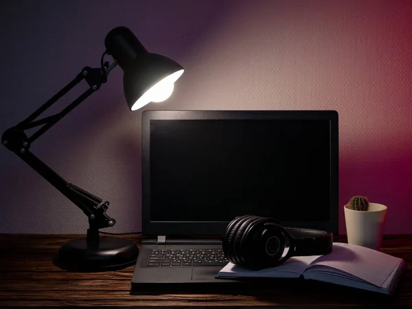 Laptop, headphones, table lamp, notebook, phone and flower at workplace. Distance work concept in quarantine. Modern freelance. Distance communication. Work in the evening.