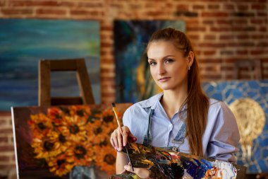 Sanat atölyesinde genç, gülümseyen kadın sanatçının portresi. Resim fırçası tutan bir ressam. Yaratıcı süreç, rahatlama, rahatlama, hobi, stres yönetimi