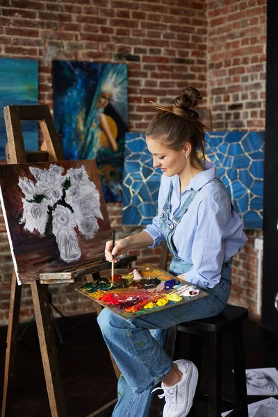 Genç ve güzel kadın ressam, yağlı boya tabloları ve sanat fırçasıyla tuval üzerinde ayçiçeğiyle resim yapıyor. Ressam sanat stüdyosunda sanat eseri yaratıyor. Rahatlama, rahatlama, hobi, stres yönetimi.