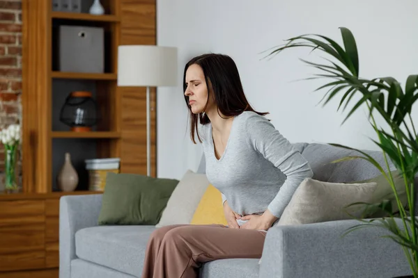 Sick young woman suffering from menstrual pain. Woman with hands squeezing belly having painful stomach ache or period cramps sitting on sofa, Abdominal pain, gastritis, and painful periods concept.