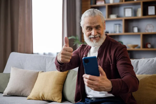 Smiling older man sitting on couch giving thumb up while playing computer game, looking at camera, smiling