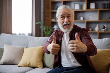 Kanepede oturmuş gülümseyen yaşlı bir adam bilgisayar oyunu oynarken başparmağını bırakıyor, kameraya bakıyor, gülümsüyor.