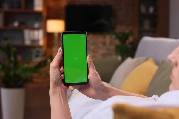 Over shoulder view of young woman holding smartphone with green mock-up screen horizontal mode. Woman lying on couch at home in evening, watching content videos blogs, news or films.Cinematic lighting