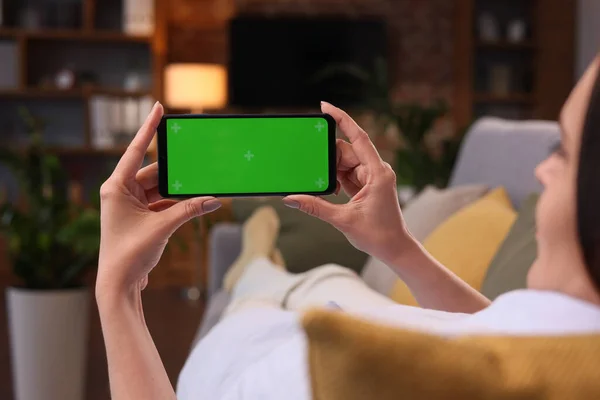 Over shoulder view of young woman holding smartphone with green mock-up screen horizontal mode.Woman lying on couch at home in evening, watching content videos blogs, news or films.Cinematic lighting