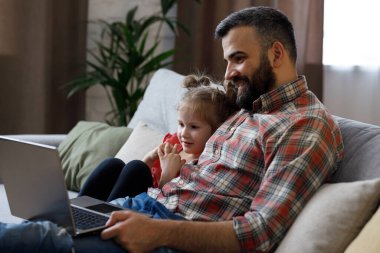 Baba ve kızı komik videolar izliyor, dizüstü bilgisayarda gülen yüzle televizyon izliyorlar. Mutlu bir aile bilgisayarda dinleniyor, hafta sonları evde birlikte vakit geçiriyorlar. Babalar Günü.