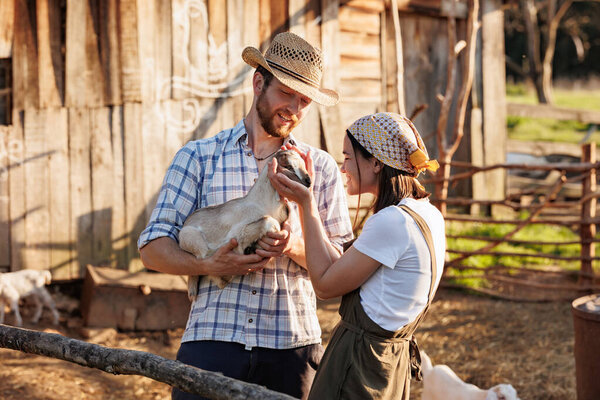 Портрет молодой кавказской счастливой фермерской пары, держащей на руках маленького козла. Семейный бизнес на экологической ферме. Здоровое питание. Промышленное производство молочных продуктов из козьего молока. Скотоводство