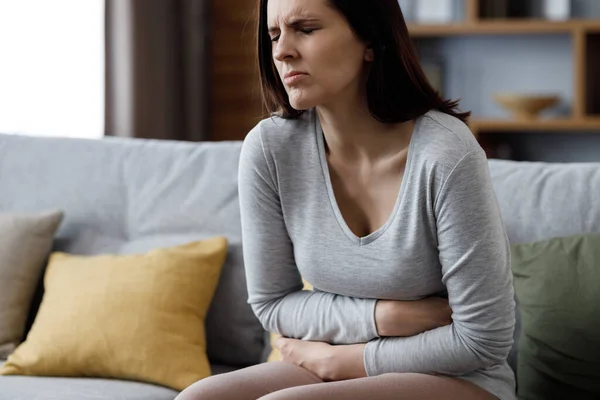 Sick young woman suffering from menstrual pain. Woman with hands squeezing belly having painful stomach ache or period cramps sitting on sofa, Abdominal pain, gastritis, and painful periods concept.