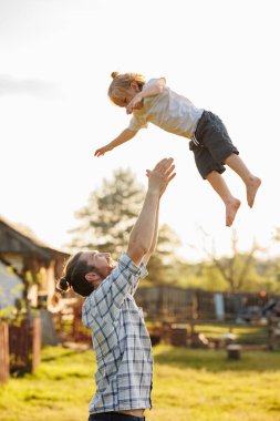 Sıcak ve güneşli bir günde babasıyla evcil bahçesinde eğlenen küçük mutlu bir çocuk. Baba çocuğunu havaya fırlatıyor. Hafta sonları birlikte kırsalda vakit geçiriyoruz. Babamla hafta sonu. Mutlu bir aile..