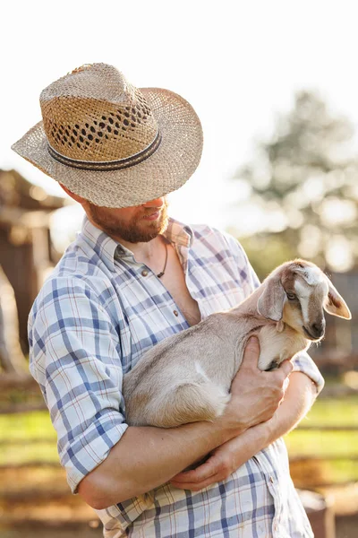 Portrait of young Caucasian bearded male farmer in straw hat holding a little goat in his arms. Happy rancherman at the eco farm. Industrial production of goat milk dairy products. Cattle farming