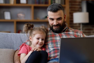 Baba ve kızı komik videolar izliyor, dizüstü bilgisayarda gülen yüzle televizyon izliyorlar. Mutlu bir aile bilgisayarda dinleniyor, hafta sonları evde birlikte vakit geçiriyorlar. Babalar Günü.