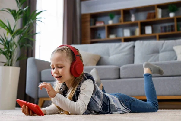 Little girl wearing red earphones lying on the carpet at home. Cute smiling child holding smartphone, watching cartoons, browsing internet or watching video in social media, playing online games.