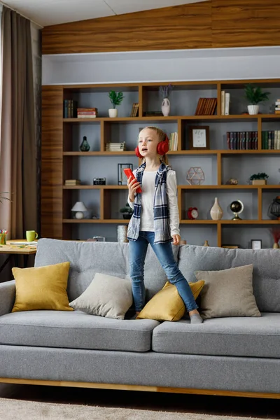 Kırmızı kulaklık takan, dans eden ve şarkı söyleyen mutlu küçük kız akıllı telefonu mikrofon gibi kullanıyor. Şirin çocuk, koça atlarken eğleniyor. Ev tatili, mutlu çocuk. Dikkat eksikliği hiperaktivite bozukluğu