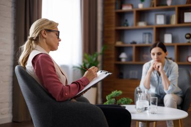 Upset young woman has counseling session with female psychologist in the office, telling her about problems. Psychologist taking notes on clipboard. Psychology, mental therapy, mental health.