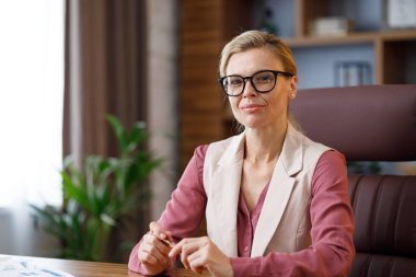 Portrait of beautiful blondie woman looking at camera and smiling. Adult stylish confident businesswoman wearing eyeglasses sitting at workplace in office. Portrait of modern successful female leader.