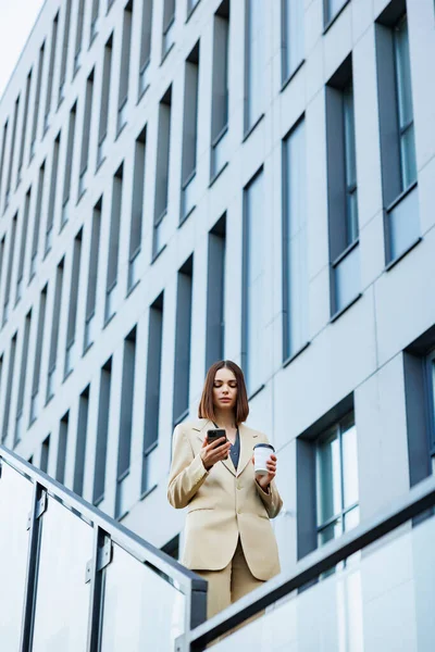 A young brunette girl against the backdrop of a business center, office center. Portrait of a successful startup. Smartphone, coffee casual suit