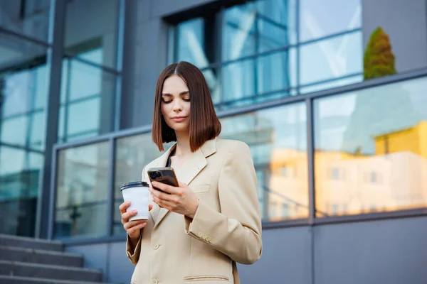 A young brunette girl against the backdrop of a business center, office center. Portrait of a successful startup. Smartphone, coffee casual suit