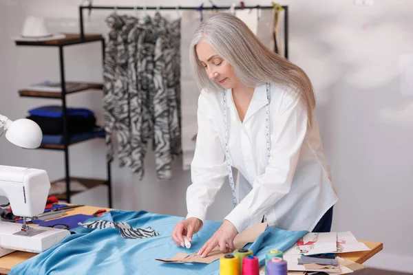 Woman with gray hair, fashion designer, stylist. Stands at the sewing table, marking the fabric before sewing