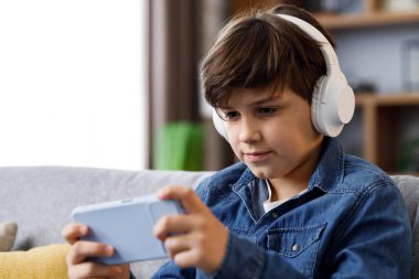 Portrait of young cute boy wearing earphones and holding smartphone sitting at sofa. Teenager at home texting messages, browsing internet, watching video in social media,playing games on mobile phone.