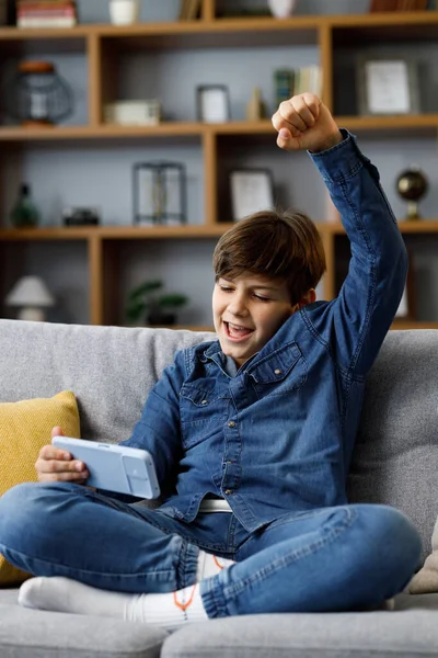 Teens and gaming addiction. Young boy playing video game on smartphone sitting on sofa at home. Cute teenager celebrating victory in online game on mobile phone. Spending time at home, leisure, relax.
