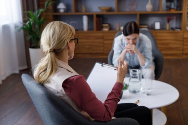 Over shoulder view of female psychologist sitting in armchair, talking with upset woman patient. Psychologist taking notes on clipboard. Psychology, mental therapy, mental health, therapy session.