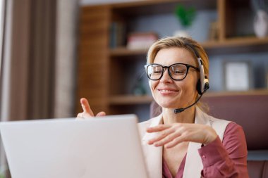 Ofiste oturmuş dizüstü bilgisayarla konuşan kulaklık takan gülümseyen iş kadını. Vlog kaydı, çevrimiçi video konferansı, video görüşmesi, online eğitim. İş görüşmesi, müşteri desteği.