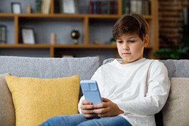 Portrait of young cute boy holding smartphone sitting at sofa. Teenager at home choosing favorite music, texting messages, browsing internet, watching video in social media, playing online games