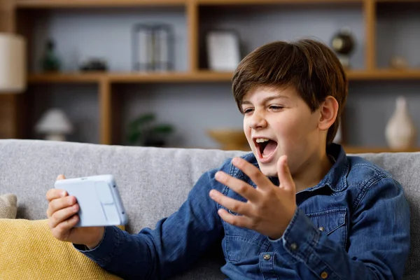 Young boy reacts emotionally to what is happening on the smartphone screen. Teenager spending time at home with digital gadget. Adolescence and puberty.